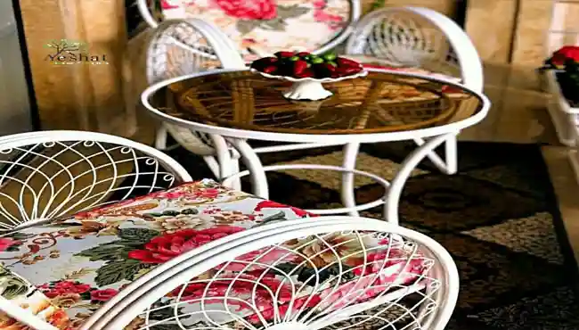 ست میز و صندلی باغی فلزی سفید سه‌نفره با تشک صندلی باغی گل‌دار صورتی و کرم، طرح گل‌های رز بزرگ، همراه میز شیشه‌ای قهوه‌ای و ظرف میوه روی آن، مناسب فضای داخلی یا تراس| گالری نشاط